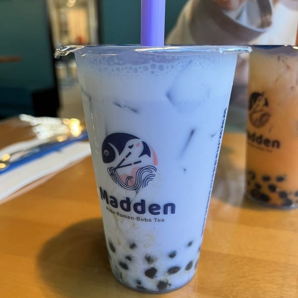 A Madden taro boba tea with a purple straw and another boba tea in the background.